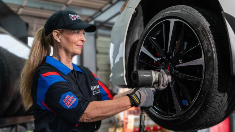 A mechanic wearing safety glasses and gloves uses a power tool to work on a car tire in an auto repair shop.