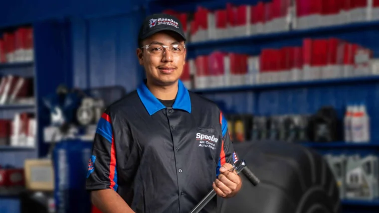 A mechanic wearing a uniform, safety glasses, and a cap stands in an auto repair shop holding a large wrench. Shelves with red and white containers are in the background.