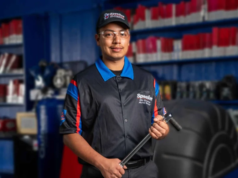 A mechanic wearing a uniform, safety glasses, and a cap stands in an auto repair shop holding a large wrench. Shelves with red and white containers are in the background.