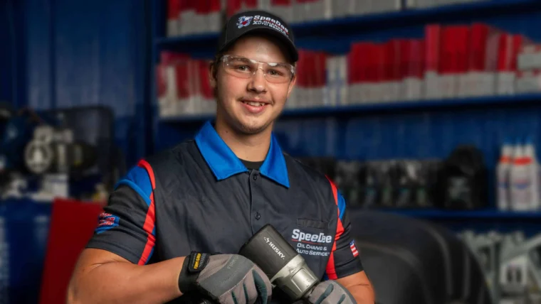 A mechanic wearing a Speedee uniform, gloves, and safety glasses holds a pneumatic tool in an automotive workshop.