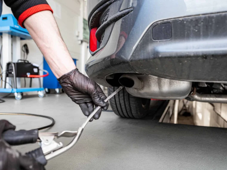 A person wearing black gloves inserts a probe into a car’s exhaust pipe, likely conducting an emissions test in a garage setting.