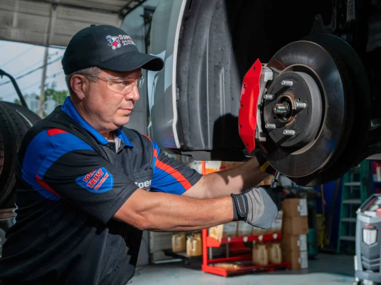 An auto mechanic in uniform and safety glasses works on the brake assembly of a car in a garage.