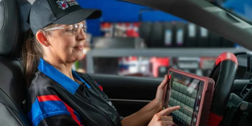 A mechanic in uniform sits in a car using a touchscreen diagnostic tool, with shelves of equipment visible in the background.