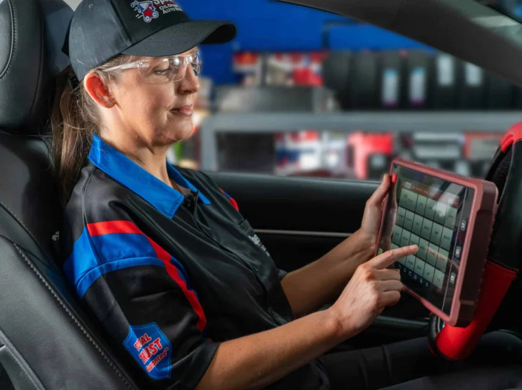 A mechanic in uniform sits in a car using a touchscreen diagnostic tool, with shelves of equipment visible in the background.