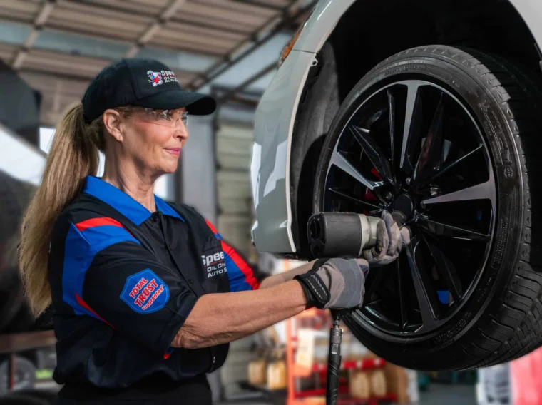 A mechanic wearing safety glasses and gloves uses a power tool to work on a car's wheel in an auto repair shop.