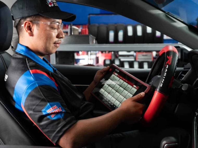 A mechanic sits in a car using a diagnostic tablet, wearing safety glasses and a uniform with patches on the sleeve while performing Tune Up Services.