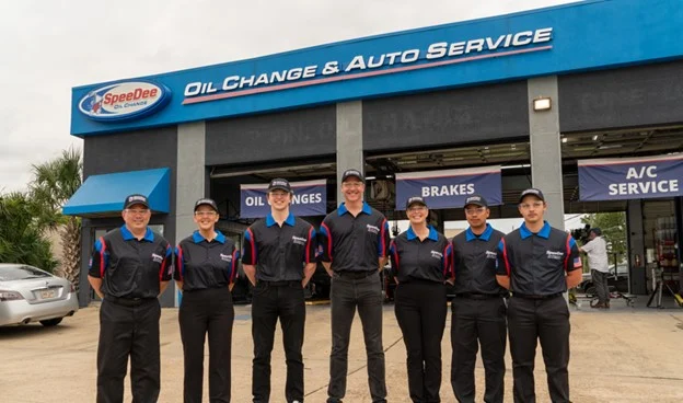 Seven employees in uniform stand in front of a SpeeDee Oil Change & Auto Service shop with service signs above the garage doors.