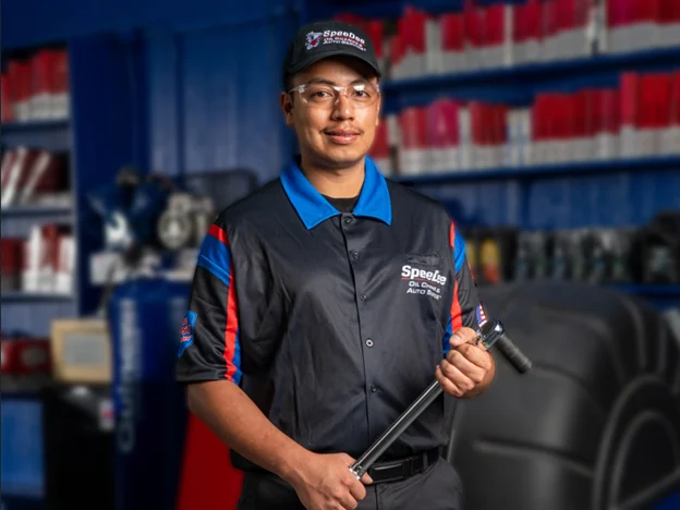 A mechanic in a black Speedee Auto Service uniform holds a tool in an auto shop with shelves of red and white boxes in the background.