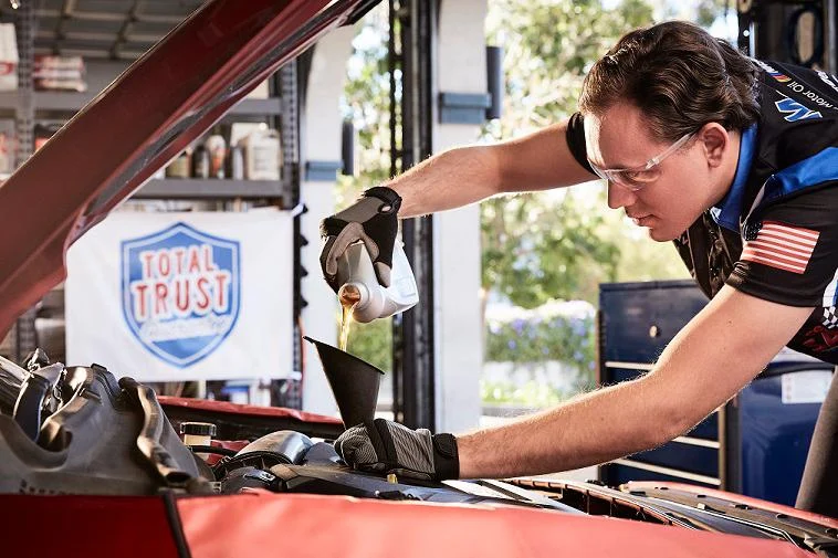 A mechanic wearing gloves and safety glasses pours engine oil into a car using a funnel, with a "Total Trust" sign visible in the background.