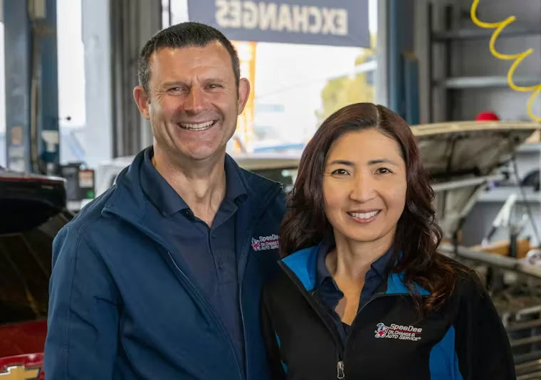 Two people wearing uniforms stand and smile inside an auto repair shop, with cars and repair equipment visible in the background.