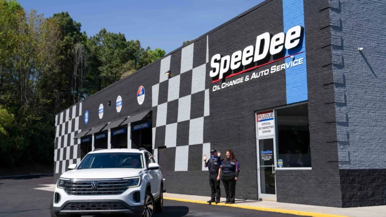 A white SUV is parked outside a SpeeDee Oil Change & Auto Service building, with two employees standing near the entrance.