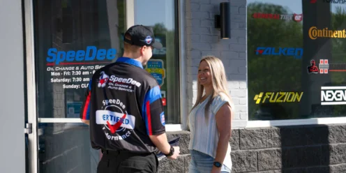A SpeeDee employee talks to a woman outside an auto service shop, with business hours and tire brand logos visible on the window.