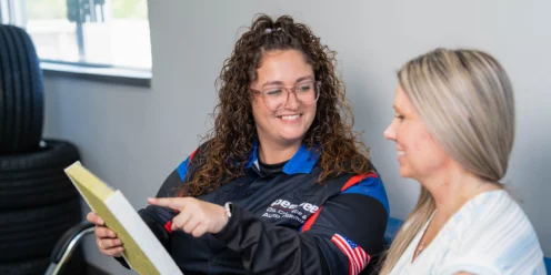 An auto service employee holding a clipboard talks with a smiling customer in a waiting area with tires stacked in the background.