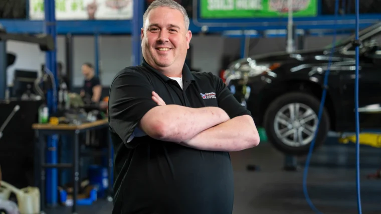 A man in a black polo shirt stands with arms crossed in an auto repair shop; a car is lifted on a hydraulic lift in the background.