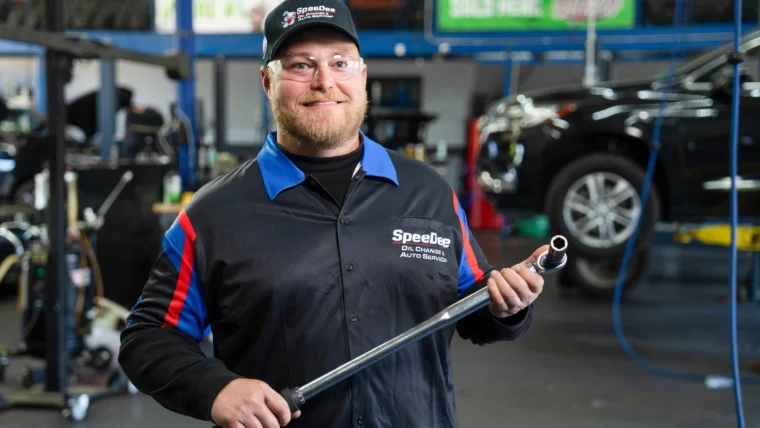 A mechanic in a Speedee Oil Change uniform stands in an auto repair shop holding a large wrench, with vehicles and equipment in the background.