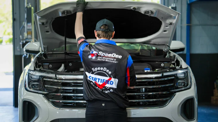 A mechanic wearing a "Speedee Certified" uniform inspects the engine of a white SUV with the hood open inside an auto service garage.