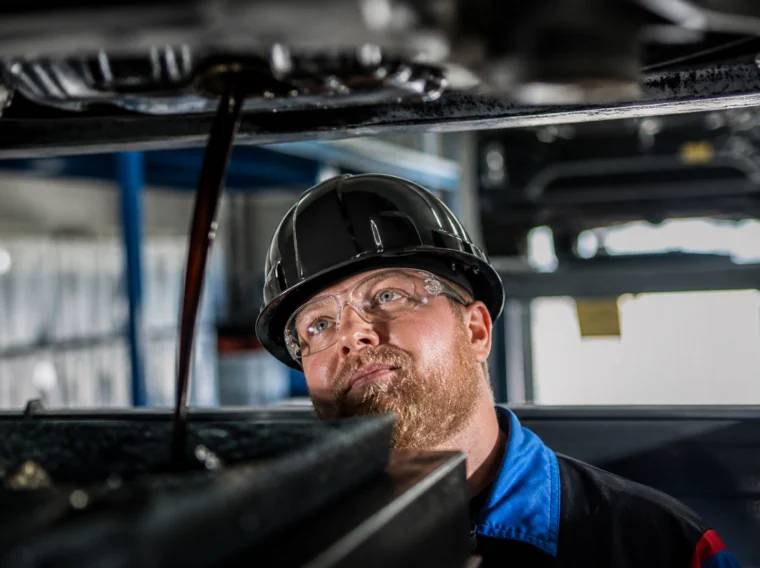 Mechanic in hard hat and safety glasses watches dark liquid draining from under a car.