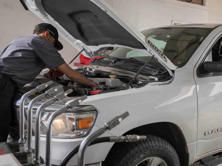 A mechanic performs battery services while repairing the engine of a white Ram truck with its hood open, as a person sits in the driver's seat inside a garage.